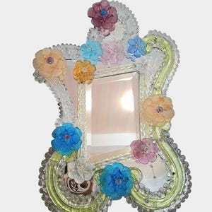 May include: A decorative mirror with a clear glass frame, adorned with colourful, translucent flowers in shades of pink, blue, yellow, and orange. The frame has a wavy, ornate design with a light green border and clear beaded accents.