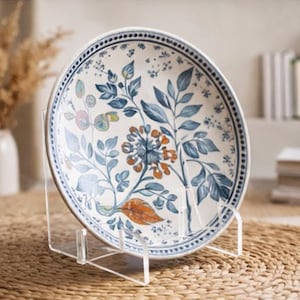 May include: A decorative ceramic plate with a floral design in blue, orange, and green. The plate is displayed on a clear acrylic stand, showcasing the intricate botanical pattern. The plate has a dark blue border.