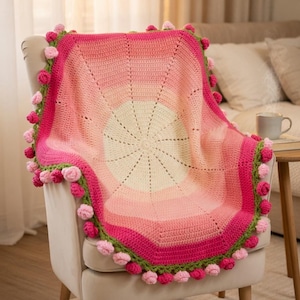 Könnte beinhalten: Eine handgehäkelte Decke in Rosa- und Cremetönen, die über einem Stuhl drapiert ist. Die Decke hat ein kreisförmiges Design mit Farbverlauf und ist mit rosa Pompons und grünen Kanten versehen. Ein gemütliches Wohnaccessoire.