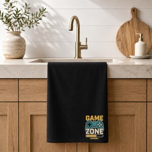 Peut inclure: Un torchon de cuisine rectangulaire noir avec une impression graphique dans la partie inf&eacute;rieure. Le motif comprend le texte jaune "GAME ZONE" au-dessus d'une ic&ocirc;ne de manette bleue et une barre de chargement jaune avec le texte "LOADING..."