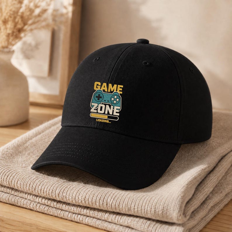 Peut inclure: Une casquette de baseball noire avec une impression graphique sur le devant. Le motif comprend une ic&ocirc;ne de manette de jeu vid&eacute;o bleu sarcelle au-dessus du texte GAME ZONE en jaune et blanc, avec une barre de chargement jaune et le mot LOADING.