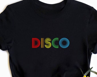 May include: Black t-shirt featuring the word "DISCO" in rainbow colours. The text has a retro design. The t-shirt is knotted at the waist. Yellow flowers are in the lower right corner.