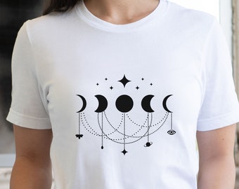 May include: White t-shirt with a black graphic design of a crescent moon, full moon, and stars connected by chains. The design also includes a small bee, a teardrop, a planet, and an eye.