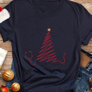 May include: Navy blue t-shirt with a red Christmas tree design and stethoscope outline. The tree has a gold star on top. The shirt is folded on a wooden surface with Christmas baubles and a Santa hat.