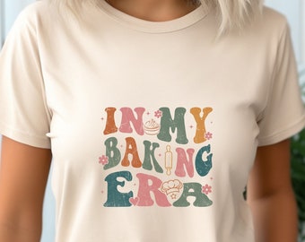 以下が含まれることがあります： ベージュのTシャツに、レトロなスタイルの「In My Baking Era」というグラフィックがプリントされています。グラフィックには、麺棒、シェフの帽子、カップケーキが描かれています。