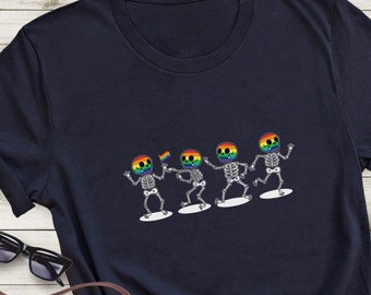 Puede incluir: Camiseta azul marino con un diseño de cuatro esqueletos bailando con calaveras de arcoíris y una bandera arcoíris. Los esqueletos están sobre plataformas blancas. El diseño celebra el orgullo LGBTQ+.