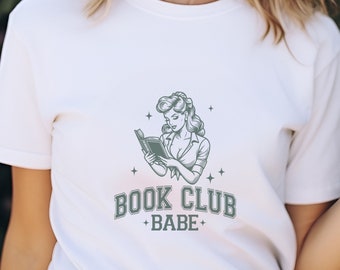 May include: White t-shirt featuring a vintage-style illustration of a woman reading a book, with the text "BOOK CLUB + BABE+" in a green-gray color. The design includes stars and a retro aesthetic.