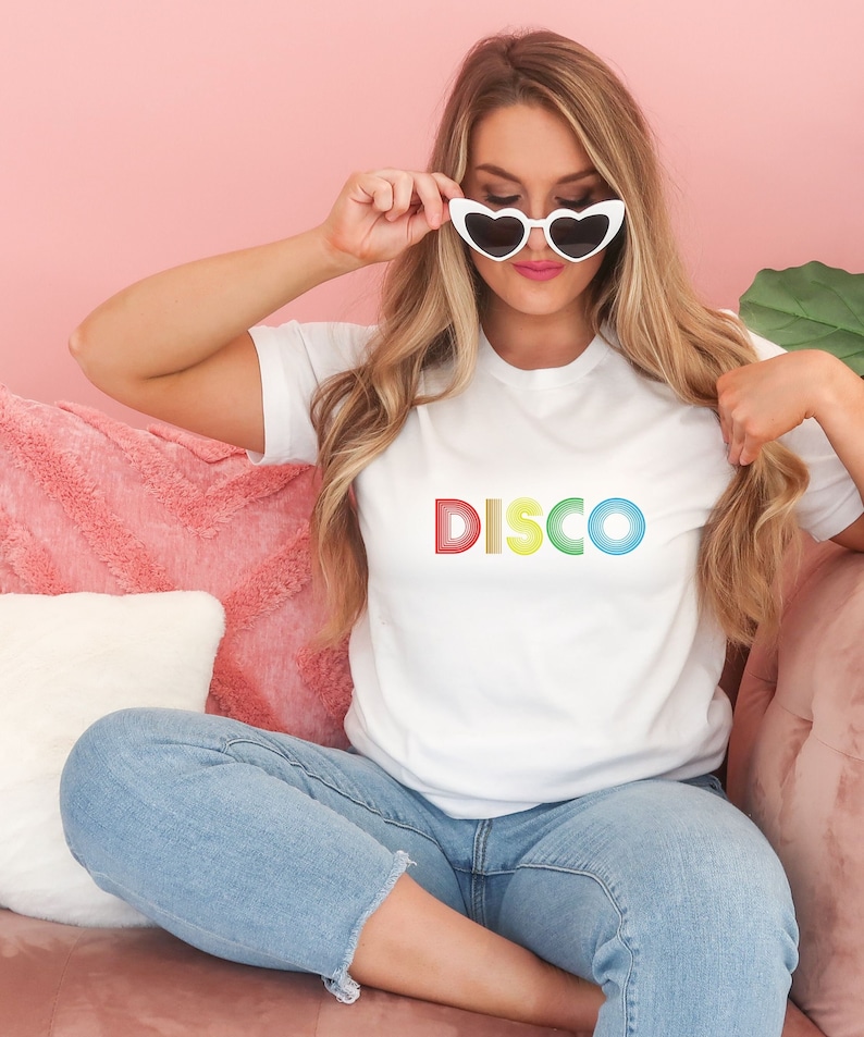 May include: A white t-shirt with the word "DISCO" printed in rainbow colors. The letters are outlined in black and have a retro, 70s style.