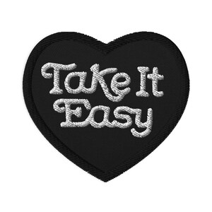 May include: A black heart-shaped embroidered patch with the words "Take It Easy" in white, cursive lettering. The patch has a stitched border and a layered design, suitable for clothing or accessories.