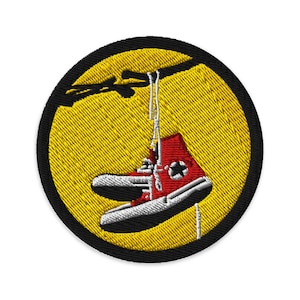 Peut inclure: Écusson brodé représentant une paire de baskets montantes rouges suspendues à une branche. Les baskets ont des semelles et des lacets blancs, avec une étoile noire sur le côté. L'écusson a un fond jaune et une bordure noire.