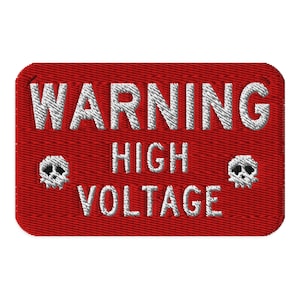 Puede incluir: Parche rojo bordado con las palabras "WARNING HIGH VOLTAGE" en hilo blanco. Dos símbolos de calavera y huesos blancos flanquean el texto. El parche tiene forma rectangular con esquinas redondeadas.