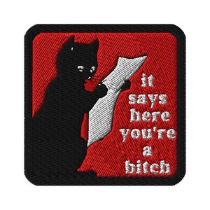 May include: Embroidered patch featuring a black cat silhouette reading a scroll. The patch has a red background and the text "it says here you're a bitch" in white. The patch has a black border.