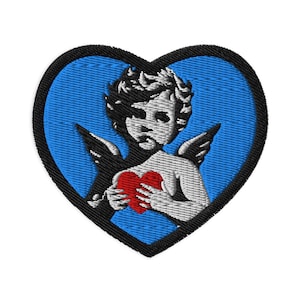 May include: Embroidered patch in the shape of a heart with a blue background and a black border. The patch features a cherub holding a red heart, with black wings and detailed shading.