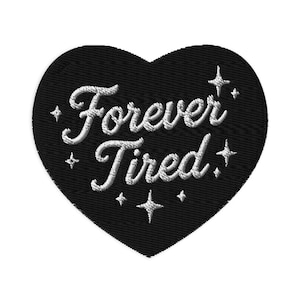 Op de afbeelding: Een hartvormige zwarte geborduurde patch met de witte tekst Forever Tired in een cursief lettertype. Het ontwerp is versierd met verschillende kleine witte vierpuntige sterpictogrammen die rond de tekst op de zwarte stoffen ondergrond staan.