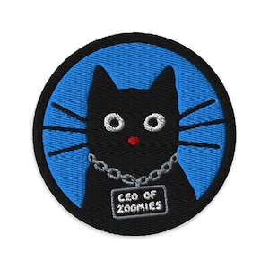 May include: A circular embroidered patch featuring a black cat with white eyes, a small red nose, and long black whiskers. The cat wears a silver-colored chain necklace with a rectangular tag that reads CEO OF ZOOMIES against a bright blue background.