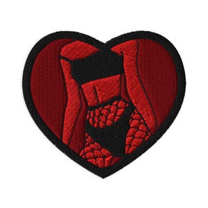 May include: Embroidered heart-shaped patch with a black border. The heart features a red design with a central figure and flowing elements. The patch is likely for clothing or accessories.