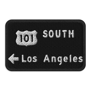 Peut inclure: Écusson noir brodé avec du texte blanc et un panneau routier blanc. Le texte indique "101 SOUTH" et "Los Angeles" avec une flèche pointant vers la gauche. L'écusson a une bordure noire.
