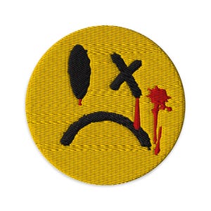 May include: A circular embroidered patch featuring a yellow background with a black sad face design. The face has one solid black oval eye, one black X-shaped eye, and a curved black mouth. Red embroidered drips extend from the eyes and mouth.