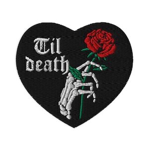 May include: Heart-shaped embroidered patch with the words "Til death" in white. A skeleton hand holds a red rose with green leaves, set against a black backdrop.