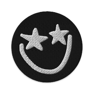 Op de afbeelding: Ronde, zwarte geborduurde patch met een lachend gezicht. De ogen zijn stervormig en de mond is gebogen, allemaal in witte stiksels. Geschikt om op kleding of accessoires te naaien of te strijken.