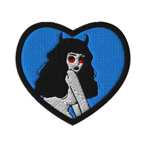 May include: Embroidered patch featuring a stylized illustration of a figure with black hair, red eyes, and horns, set against a blue background within a black-outlined heart shape. The figure is in a white and gray color scheme.