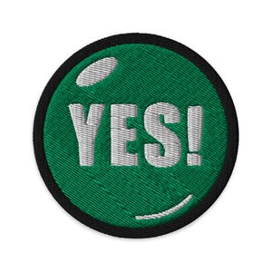 Puede incluir: Parche bordado con fondo verde y la palabra "YES!" en blanco. El parche tiene un borde negro y un aspecto texturizado. Un diseño curvo blanco en la parte inferior.