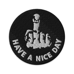 May include: A circular black embroidered patch featuring a white stitched graphic of a hand making a middle finger gesture. The text "HAVE A NICE DAY" is embroidered in white capital letters in a curved arc along the bottom of the patch.