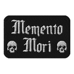 Könnte beinhalten: Ein rechteckiger, schwarz bestickter Aufnäher mit dem weißen Text Memento Mori in einer gotischen Schriftart. Das Design enthält ein kleines weißes Totenkopf-Symbol links und rechts neben dem Wort Mori, alles auf einem schwarzen Stoffhintergrund.