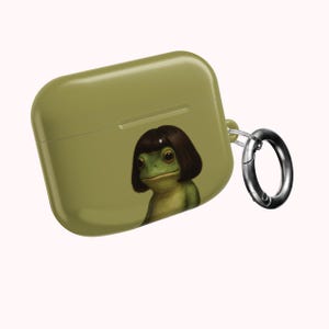 Puede incluir: Funda para AirPod de color verde oliva con una ilustración caprichosa de una rana con un corte de pelo bob castaño oscuro. La funda tiene un mosquetón plateado. La rana tiene ojos grandes y expresivos y una expresión serena.