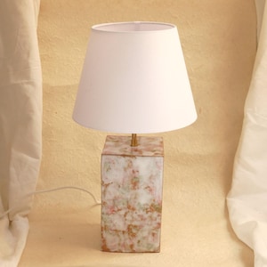May include: A decorative table lamp with a white lampshade and a rectangular base. The base features a marbled design with shades of white, pink, green, and gold. The lamp has a brass stem and a white cord.