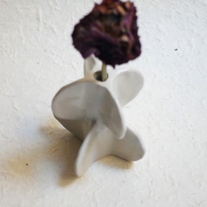 May include: A white ceramic vase with a unique, geometric design. The vase has a spiral shape with multiple layers of petals. A dried purple rose is placed in the vase.