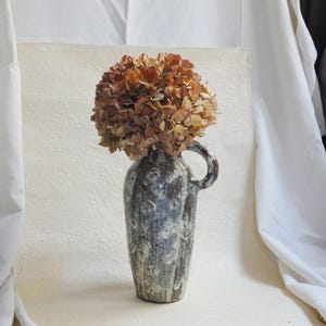 May include: A ceramic vase with a unique, textured glaze in shades of blue and white. The vase has a rounded body and a handle on the side. It is filled with dried hydrangeas in shades of brown and pink.