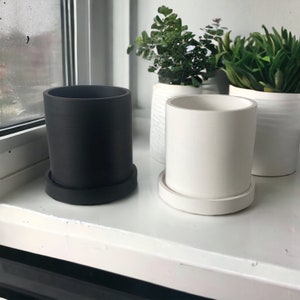May include: Two cylindrical plant pots, one black and one white, each on a matching saucer. The pots are on a white windowsill, with other white pots holding greenery in the background. The pots are designed for indoor use.