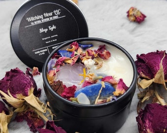 May include: A black candle tin with the text "Witching Hour LLC All Natural Intention Soy Candle Sleep Tight Bergamot, Jasmine, Neroli & Musk Hand-Poured 1.2 oz" on the lid. The candle is topped with dried rose petals, crystals, and a clear wax layer.