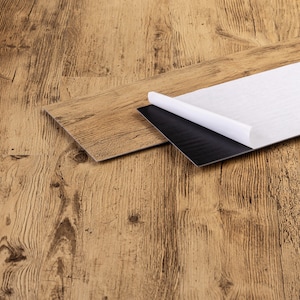 Könnte beinhalten: Nahaufnahme von Bodenbelagsplanken in Holzoptik. Eine Planke ist teilweise abgezogen und zeigt eine schwarze Klebeschicht und eine weiße Rückseite. Die Planken sind hellbraun mit Holzmaserung, geeignet für Heimwerkerprojekte.