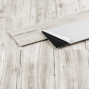 Może przedstawiać: Zbliżenie jasnoszarych paneli podłogowych o wyglądzie drewna. Jedna deska jest częściowo odklejona, odsłaniając czarny klejący spód. Podłoga ma teksturowany, postarzany wygląd z widocznym usłojeniem drewna.