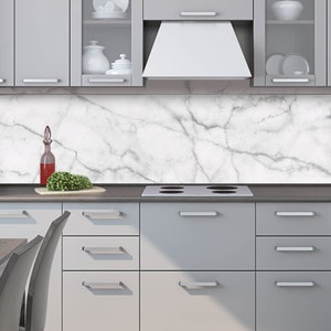 Può includere: Un paraschizzi da cucina in marmo bianco con venature grigie. Il paraschizzi è installato sopra un pensile da cucina grigio con un piano cottura in acciaio inox.