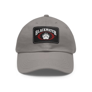 Puede incluir: Gorra de béisbol gris con visera curva. La gorra presenta un parche rectangular con la palabra "BLACKWATER" en blanco, texto arqueado sobre un óvalo rojo que contiene una huella de pata blanca.