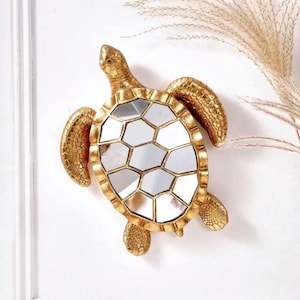 Könnte beinhalten: Ein goldfarbener, schildkrötenförmiger Wandspiegel. Der Spiegel hat ein segmentiertes, geometrisches Design und ist von einem strukturierten, goldenen Rahmen umgeben. Kopf, Flossen und Beine der Schildkröte sind ebenfalls goldfarben. Der Spiegel ist an einer weißen Wand befestigt.