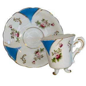 May include: A white teacup and saucer set with blue and gold accents. The cup has a unique three-legged design and a handle. Floral patterns in pink and green decorate the cup and saucer, which has a scalloped edge.