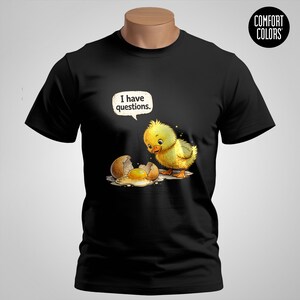 Op de afbeelding: Zwart T-shirt met een cartoonkuiken dat naar een gebroken ei en eidooier kijkt. Een tekstballon boven het kuiken luidt: "Ik heb vragen". Het shirt heeft een Comfort Colors-logo rechtsboven.