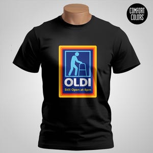 May include: Black t-shirt with a humorous graphic. The graphic features a blue stick figure using a walker inside a colorful frame, with the word "OLDI" and the text "Still Open at 8pm". The shirt has a "Comfort Colors" logo.