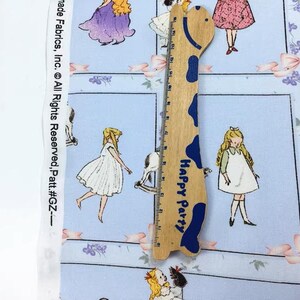 Pode incluir: Tecido azul claro com um padr&atilde;o repetido de ilustra&ccedil;&otilde;es vintage de meninas em vestidos, la&ccedil;os e detalhes florais. Uma r&eacute;gua de madeira em forma de girafa com o texto "Happy Party" est&aacute; colocada sobre o tecido.