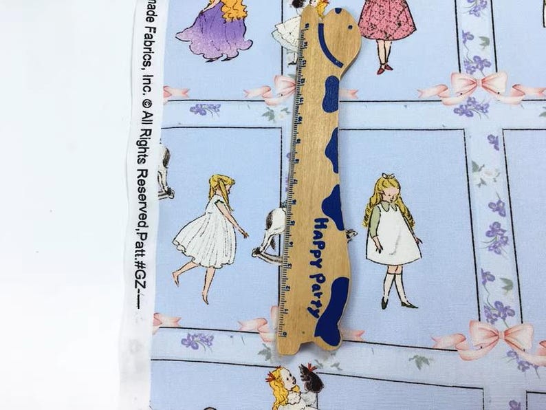Pode incluir: Tecido azul claro com um padr&atilde;o repetido de ilustra&ccedil;&otilde;es vintage de meninas em vestidos, la&ccedil;os e detalhes florais. Uma r&eacute;gua de madeira em forma de girafa com o texto "Happy Party" est&aacute; colocada sobre o tecido.