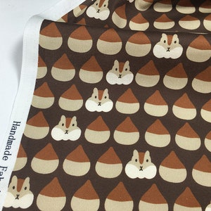 Op de afbeelding: Bruine stof met een herhalend patroon van bruine en beige eikels en witte eekhoorns. De stof heeft een witte rand met de tekst "Handmade Fabrics" erop gedrukt.