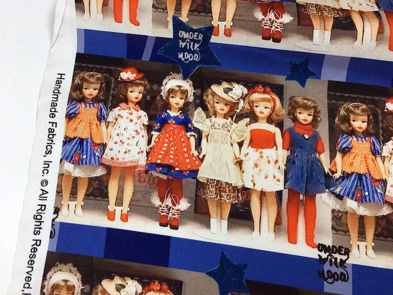 Op de afbeelding: Een kleurrijke stof met een herhalend patroon van poppen die verschillende jurken en hoeden dragen. De stof heeft een blauwe en witte achtergrond met sterren en de tekst "Under Milk Wood" die zich over de hele stof herhaalt.