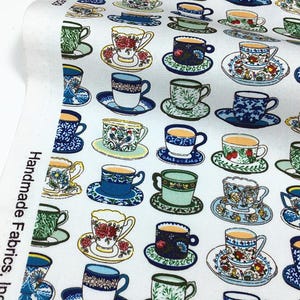 Può includere: Tessuto bianco stampato con un motivo ripetuto di tazze e piattini colorati. Le tazze presentano vari disegni floreali e geometrici nelle tonalità del blu, verde, giallo e rosso. Il tessuto è etichettato "Handmade Fabrics, Inc."