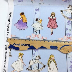 Pode incluir: Tecido azul claro com um padr&atilde;o repetido de ilustra&ccedil;&otilde;es de meninas em vestidos, cavalos de baloi&ccedil;o e la&ccedil;os. Uma r&eacute;gua de madeira com o texto "Happy Party" &eacute; colocada sobre o tecido. O tecido tem o texto "Handmade Fabrics, Inc." na lateral.