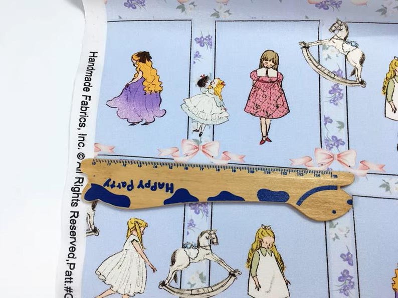 Pode incluir: Tecido azul claro com um padr&atilde;o repetido de ilustra&ccedil;&otilde;es de meninas em vestidos, cavalos de baloi&ccedil;o e la&ccedil;os. Uma r&eacute;gua de madeira com o texto "Happy Party" &eacute; colocada sobre o tecido. O tecido tem o texto "Handmade Fabrics, Inc." na lateral.
