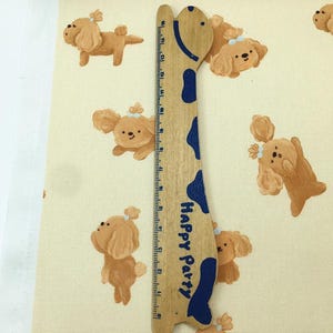 May include: A light beige fabric with a repeating pattern of cartoon dogs in various poses. A wooden ruler with blue accents and the text "Happy Party" is placed vertically on the fabric.